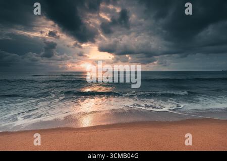 Dramatischer Sonnenuntergang am Strand über den Wellen des Ozeans mit stürmischen Wolken im Himmel Stockfoto