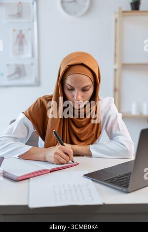 Muslimische Ärztin mit Stethoskop, die mit dem Schreiben von Patientennotizen und Diagnose mit Konzentration in der Klinik arbeitet, mit Laptop-Computer im Krankenhaus Stockfoto