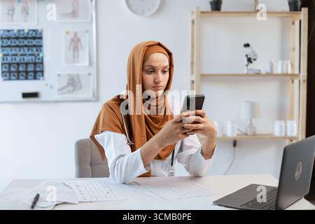 Gesundheitswesen, Medizin und Technologiekonzept. Professionelle muslimische Ärztin arbeitet in der Klinik und sendet Textnachrichten auf dem Smartphone bei der Arbeit. Kopierbereich Stockfoto