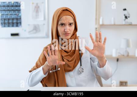 Junge muslimische professionelle Ärztin mit offenen Händen, die Stoppzeichen mit ernstem Ausdruck macht, die im Krankenhausbüro am Schreibtisch sitzt. Medicin Stockfoto