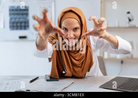 Weibliche verärgerte wütende muslimische Ärztin schreiend Hände in wütender Geste halten sitzen am Schreibtisch und machen professionelle medicare zum Klinikpatienten. Konzept von M Stockfoto