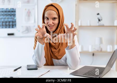 Weibliche verärgerte wütende muslimische Ärztin schreiend Hände in wütender Geste halten sitzen am Schreibtisch und machen professionelle medicare zum Klinikpatienten. Konzept von M Stockfoto