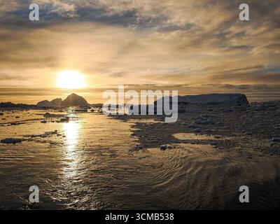 Goldener Sonnenuntergang reflektiert auf dem Polarmeer mit schwimmendem Eis und fernen Eisbergen unter dramatischen Wolken. Stockfoto