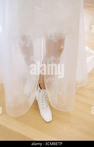 Eine Braut zeigt ihr atemberaubendes Brautkleid aus Spitze und stilvolle weiße Stiefel in einem hellen Raum. Stockfoto