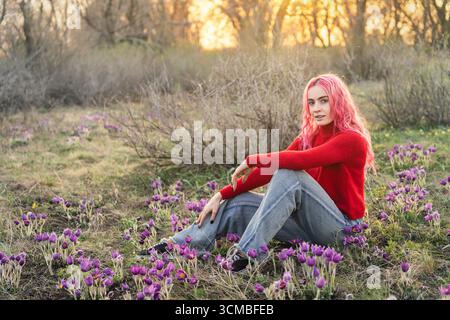 Eine junge Frau mit leuchtend rosa Haaren sitzt friedlich auf einer blühenden Wiese, umgeben von der Ruhe der Natur. Ihr knallroter Pullover bildet einen Kontrast zu bea Stockfoto