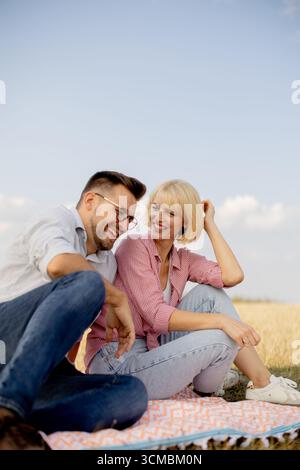 Ein fröhliches Paar sitzt zusammen auf einer Decke in einem goldenen Feld und tauscht Lächeln und Lachen unter einem klaren blauen Himmel aus. Sie wirken entspannt und glücklich Stockfoto