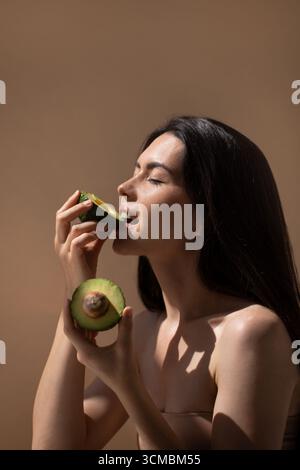 Weibliches Modell mit dunklem Haar schmeckt frische Avocado, hält die eine Hälfte, während man einen Biss von der anderen nimmt, betont Gesundheit und Wellness in einer ruhigen Stockfoto