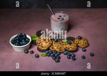 Ein lebendiger, tiefvioletter Smoothie in einem Glas mit einer Orangenscheibe, begleitet von Shortbread und Heidelbeerkeksen auf einem hellen, hellvioletten B Stockfoto