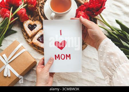Frauenhände halten eine schöne handgezeichnete Muttertagsgrußkarte mit rotem Umschlag, auf der Karte steht die Nachricht „I Love You Mom“. Stockfoto
