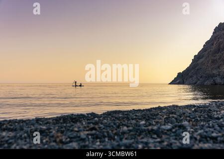 Samothraki, Griechenland – SUP-Aktivität am Kipos Beach bei Sonnenuntergang, mit zwei Personen auf einem Paddleboard über ruhigem Wasser, goldenen Reflexen und Stockfoto