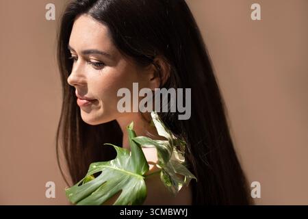 Weibliches Modell mit dunklem Haar, grünen Blättern, posiert elegant vor einem weichen Hintergrund und fängt in diesem A die Essenz der Natur und Ruhe ein Stockfoto