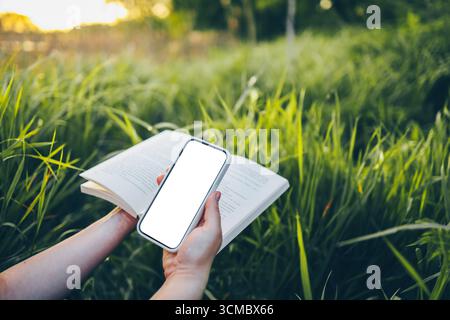 Ein offenes Buch in den Händen und ein Telefon mit einem isolierten Bildschirm, das Konzept der Natur, Ruhe und Einsamkeit. Stockfoto