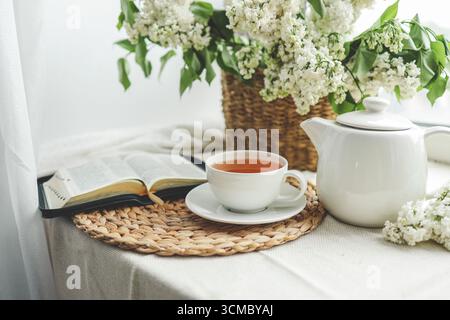 Eine Tasse Tee, ein Korb mit Flieder und eine offene Bibel. Stockfoto