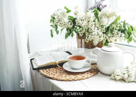 Eine Tasse Tee, ein Korb mit Flieder und eine offene Bibel. Stockfoto