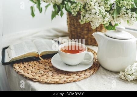 Eine Tasse Tee, ein Korb mit Flieder und eine offene Bibel. Stockfoto