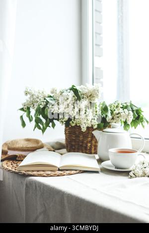 Offenes Buch, Tasse Tee und Teekanne, Korb mit weißem Flieder und Hut, Sommerkomposition. Stockfoto