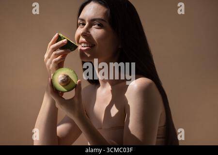 Weibliches Modell mit dunklem Haar hält Avocadohälften, lächelt und fördert gesunde Ernährung, mit sanfter Beleuchtung und einem neutralen Hintergrund, der die Natu verstärkt Stockfoto