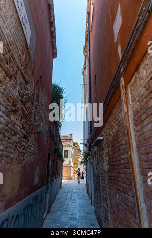 Enge Gasse, die zur sonnendurchfluteten Straße in Venedig führt. Die gemauerte Gasse führt zu einer hellen venezianischen Straße, wo Touristen an bunten Gebäuden vorbeilaufen Stockfoto