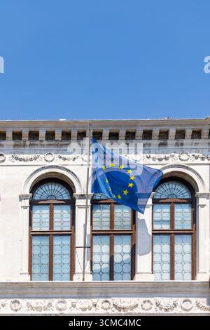 Flagge der Europäischen Union winkt auf historischem Gebäude. Die Flagge der Europäischen Union flattert vor einem reich verzierten historischen Gebäude mit Bogenfenstern und Stockfoto