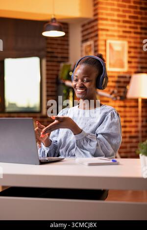 Fröhliche Frau, die virtuelle Gespräche mit Investoren und Aktionären während einer Telefonkonferenz auf einem Laptop im Home Office führt. Weibliche Führungskraft, die ein schnurloses Headset trägt, leitet Ferngespräche mit Kollegen. Stockfoto