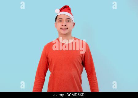Fröhliche junge Person mit weihnachtsmütze in festlicher roter Kleidung, die weihnachten auf blauem Hintergrund feiert Stockfoto