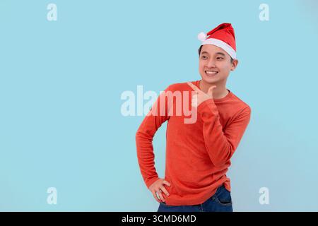 Lächelnder junger Mann im weihnachtsmannhut-Enthusiast, der auf blauen Hintergrund mit weihnachtskonzept zeigt Stockfoto