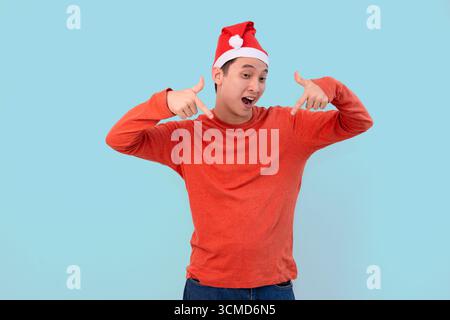 Ein fröhlicher junger Mann, der einen roten Pullover und einen Weihnachtsmann-Hut trägt, zeigt nach unten im blauen Hintergrund des weihnachtskonzepts Stockfoto