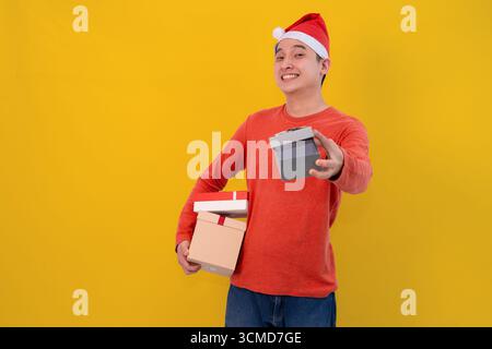 Ein junger Mann, der einen roten Weihnachtsmann-Hut trägt, bietet freudig eine verpackte Geschenkbox mit gelbem Hintergrund des weihnachtskonzepts an Stockfoto
