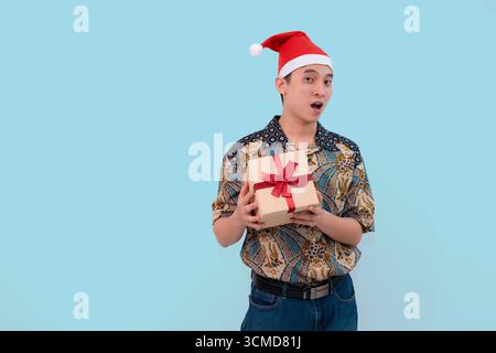 Ein fröhlicher junger Mann mit Batik und Weihnachtsmann-Hut, der eine Geschenkbox auf blauem Hintergrund hält Stockfoto