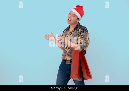 Ein fröhlicher Mann trägt Batik mit Weihnachtsmann-Hut und trägt rote Einkaufstaschen, die auf Copyspace vor einem hellblauen Hintergrund zeigen Stockfoto