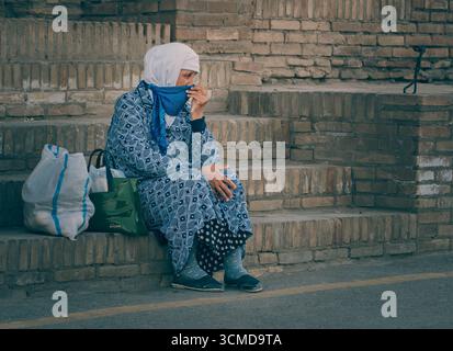 Die ältere usbekische Frau sitzt auf Ziegeltreppen in Buchara, Usbekistan. Sie zieht einen blauen Schal über das Gesicht und trägt ein gemustertes blaues Kleid. Stockfoto
