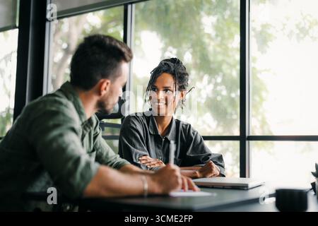 Zwei glückliche, vielseitige Mitarbeiter aus verschiedenen ethnischen Geschäftsbereichen, die im Büro des Unternehmens arbeiten und sich unterhalten Stockfoto