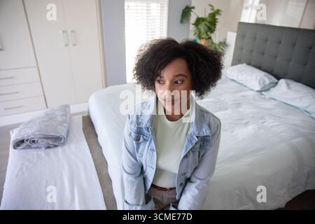Afroamerikanerin in ihren 30ern, die in einem hellen Schlafzimmer auf dem Bett sitzt und eine Jeansjacke trägt Stockfoto