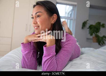 Asiatische Frau, die im Schlafzimmer mit lila Oberseite auf dem Bett liegt und auf das Fenster blickt, den Kopierraum Stockfoto