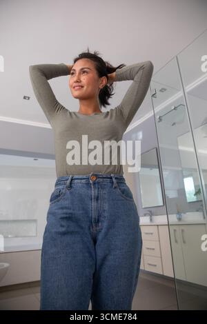 Asiatische Frau, die die Arme hebt und Haare sammelt, im Badezimmer in der Nähe einer Glasdusche und eines Kosmetikspiegels Stockfoto