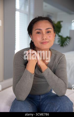 Asiatische Frau, die auf weißem Bett in einem modernen Schlafzimmer sitzt, bei Fensterläden mit Topfpflanzen dahinter Stockfoto