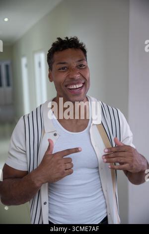 Afroamerikanischer Mann, der im Flur zu Hause steht und auf die Brust zeigt, während er den Schultergurt hält Stockfoto