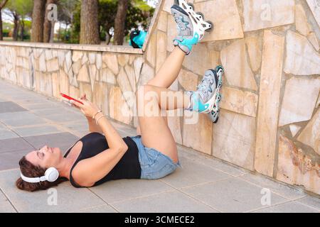 Frau, die mit Rollschuhen auf dem Boden liegt und Musik hört und Handy benutzt Stockfoto