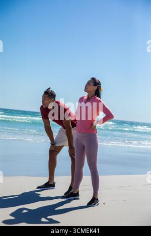 Abwechslungsreiches Paar, das Atem schnappt und sich in Laufschuhen am Sandstrand am Meeresrand ausdehnt Stockfoto