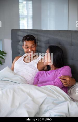 afroamerikaner und asiatisches Paar sitzen am Kopfteil und lächeln sich gegenseitig mit Bettdecke an Stockfoto
