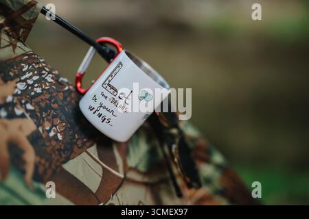 Inspirierender Becher, der an Campingausrüstung hängt, mit motivierender Typografie und Naturkulisse Stockfoto