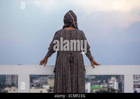 Rückansicht einer muslimischen Frau aus Bangladesch mit Blumenabaya und Hijab, die am Nachmittag auf einem Dach steht und die Stadt und die Natur betrachtet. Stockfoto