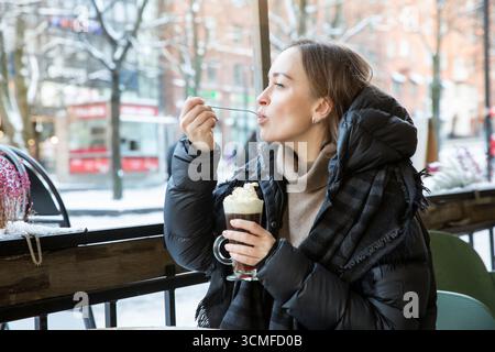 Frau mittleren Alters im Wintermantel genießt Kaffee mit Schlagsahne im verschneiten Café im Freien, Weihnachtsstimmung, gemütlicher urbaner Urlaubsstil. Stockfoto