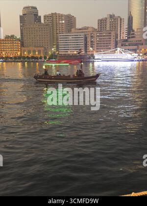 Eine Küstenszene in der Abenddämmerung mit einem Holzboot mit Passagieren und beleuchteten Gebäuden. Stockfoto