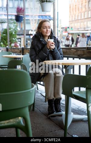 Frau mittleren Alters im Wintermantel genießt Kaffee mit Schlagsahne im verschneiten Café im Freien, Weihnachtsstimmung, gemütlicher urbaner Urlaubsstil. Stockfoto