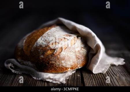 Nahaufnahme von knusprigem Sauerteigbrot mit einer rauen gebräunten Haut, die in ein Leinentuch gewickelt ist. Hausgemachte Wildhefe auf gereifter Holzoberfläche Stockfoto