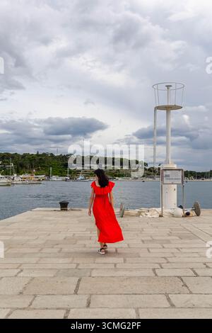 Frau in rotem Kleid, die entlang des Marina Pier läuft. Rückansicht einer Frau in fließendem rotem Kleid, die auf einem Steinpier an einem Yachthafen unter einem bewölkten Himmel läuft. Stockfoto