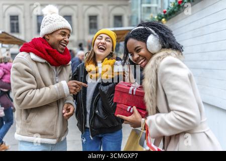Drei Freunde haben Spaß beim Austausch von weihnachtsgeschenken auf einem Wintermarkt, lachen und genießen die festliche Atmosphäre Stockfoto