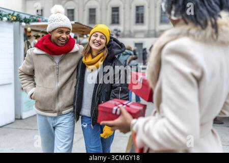 Freunde lachen und tauschen weihnachtsgeschenke auf einem Wintermarkt in der Stadt aus und genießen die festliche Atmosphäre Stockfoto