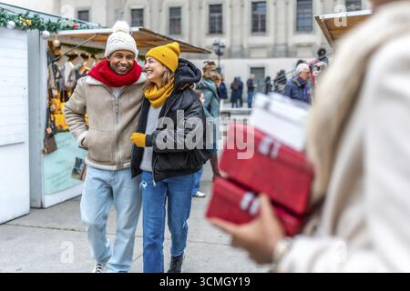 Glückliches, multiethnisches Paar, das Arm in Arm zwischen den Verkaufsständen des weihnachtsmarktes geht, eine Frau mit Geschenken Stockfoto